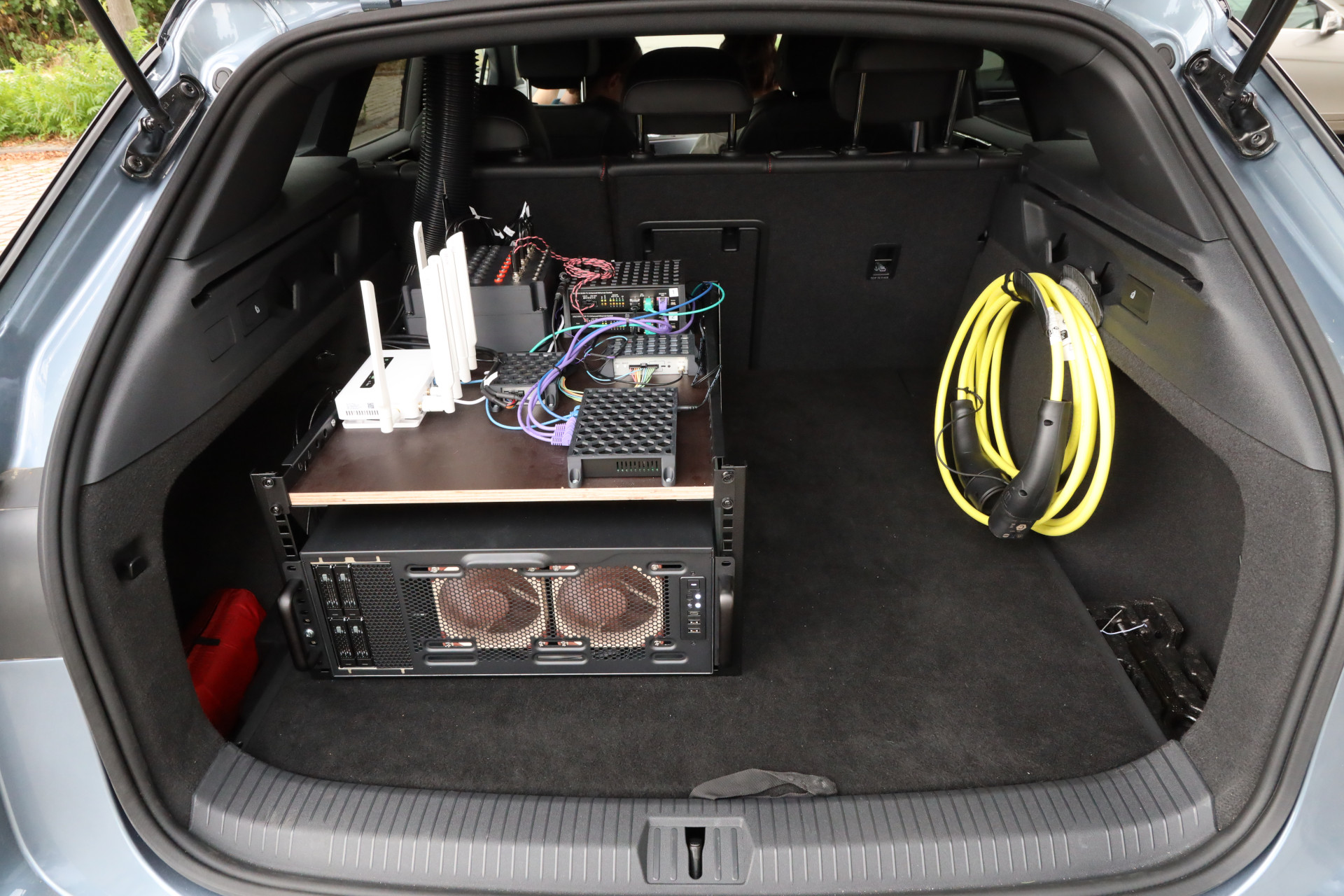 The open trunk of the research vehicle showing a 19 inch server case in a mini rack, with automotive Ethernet switches, a wifi router and cabling on a shelf above it, and a charging cable coiled to the side.