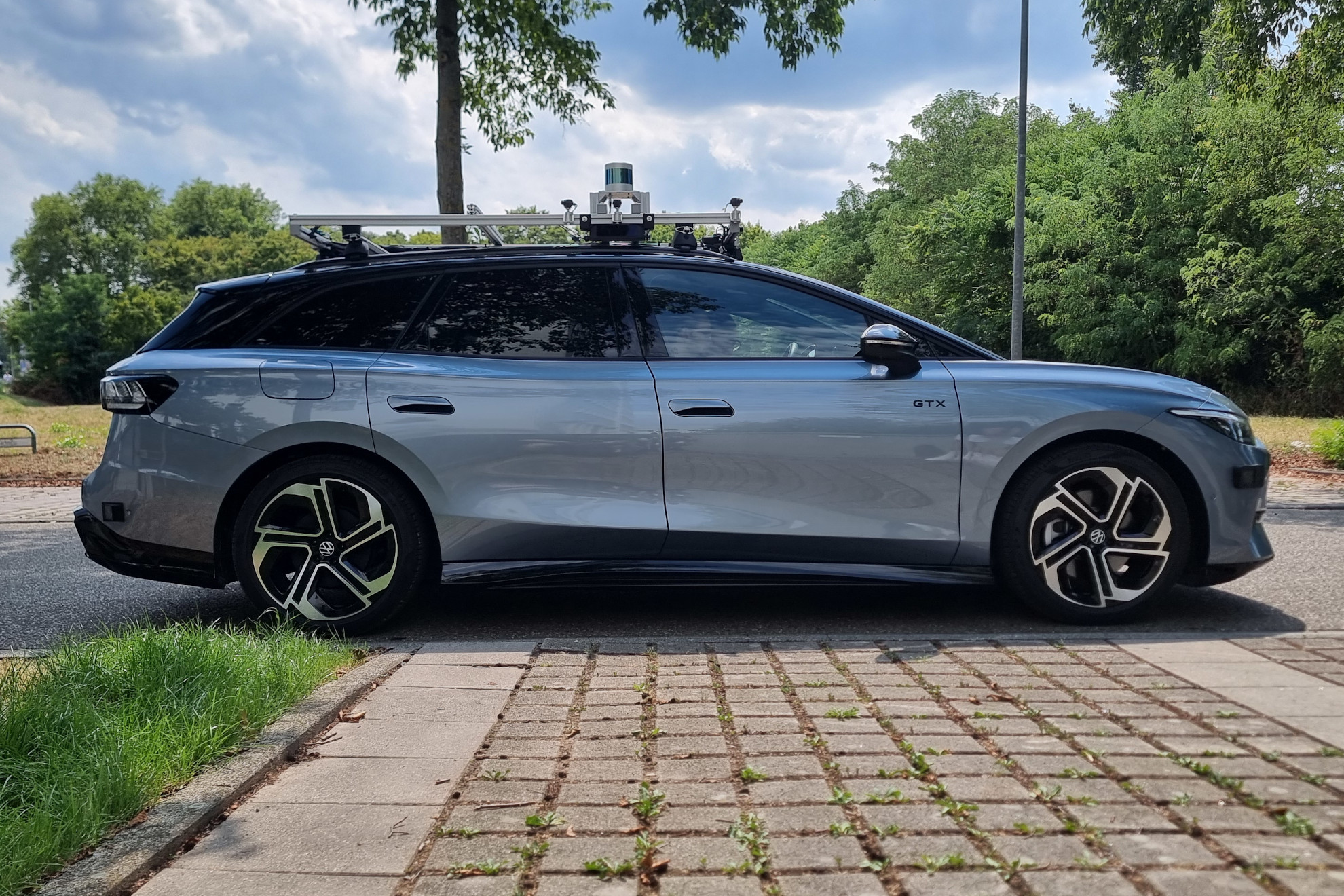 Side view of the KE:SAI research vehicle, a light blue VW ID.7 Tourer with an aluminum sensor rack on the roof carrying cameras and a 360 degree lidar on a raised tower.